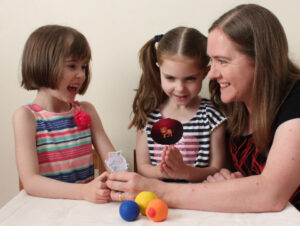 Two young girls and their mother at a table, playing with elephant and lion cut outs. Three juggling balls are on the table.