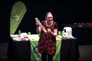 A science show presenter stands in front of a table and holds a rain stick.