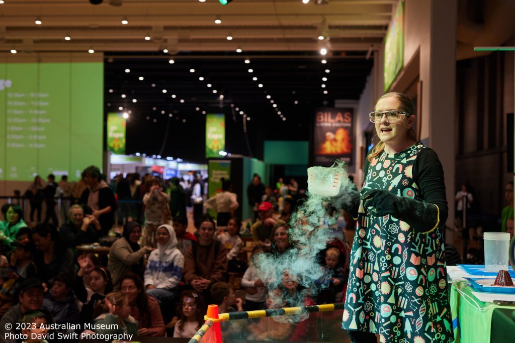 © 2023 Australian Museum - Photo David Swift Photography 
Jenny Lynch, female presenter, holding a Pyrex jug of liquid nitrogen and talking to an audience of adults and children at the Australian Museum.