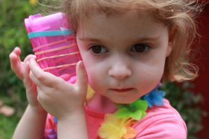 Three year old girl holding a plastic box to her ear. There are five rubber bands stretched around the box and she is plucking the rubber bands with her fingers.